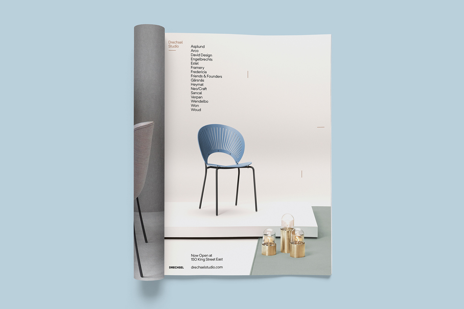 An open magazine page displays a modern blue chair on a raised white platform beside small brass lamps against a plain background