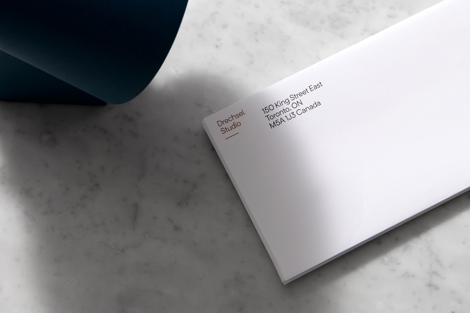 A stacked white envelope featuring the Drechsel Studio logo and address on a white marble surface next to a curved blue object.