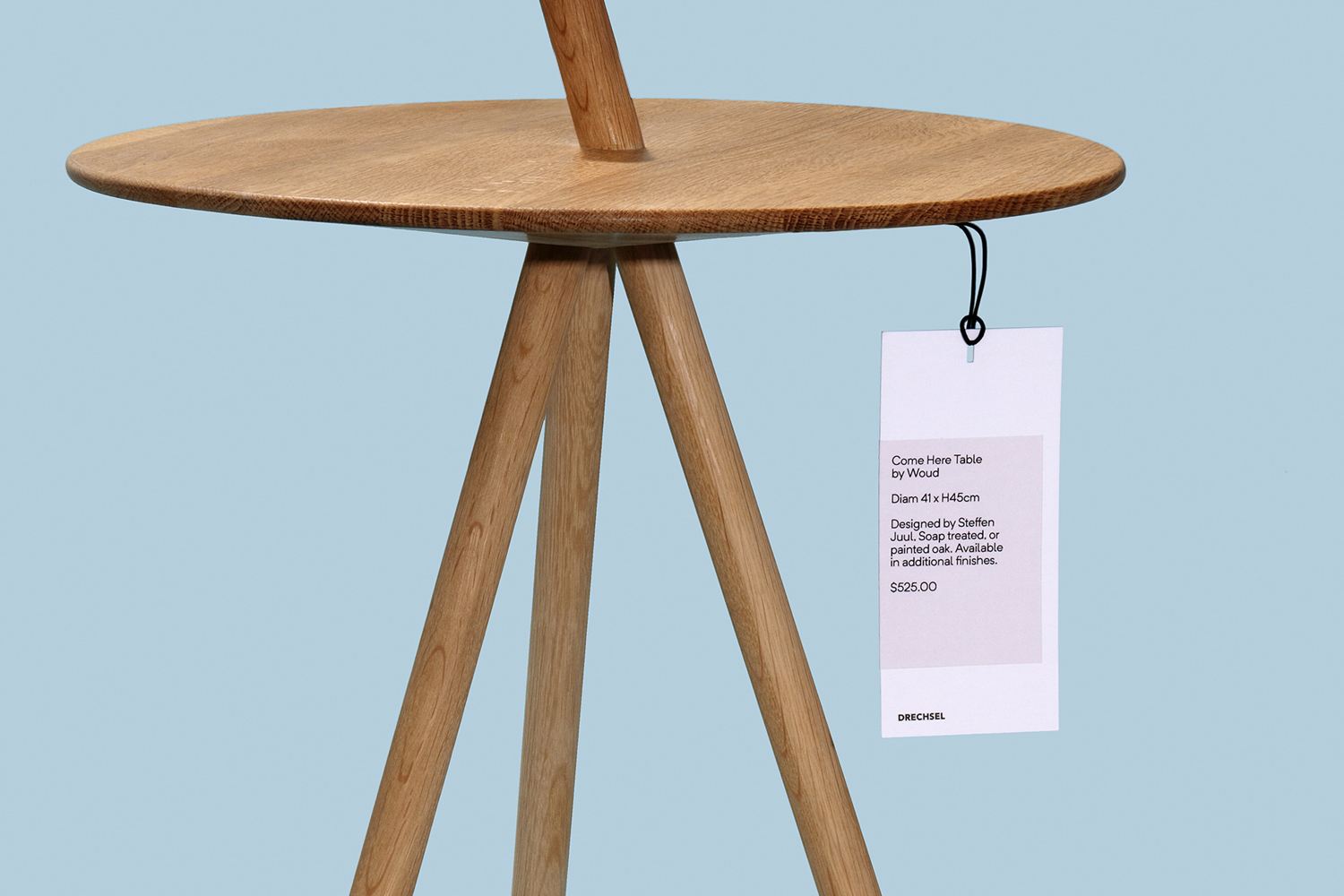 Wooden side table with three angled legs and a round top featuring a hanging product tag against a light blue background