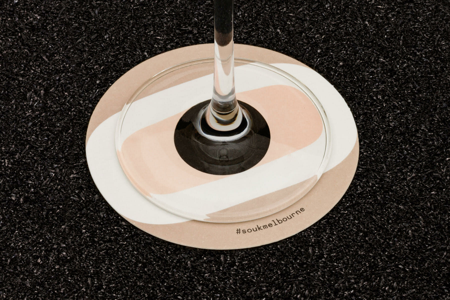Close-up of a wine glass base resting on a round patterned coaster over a dark textured surface for Souk brand identity