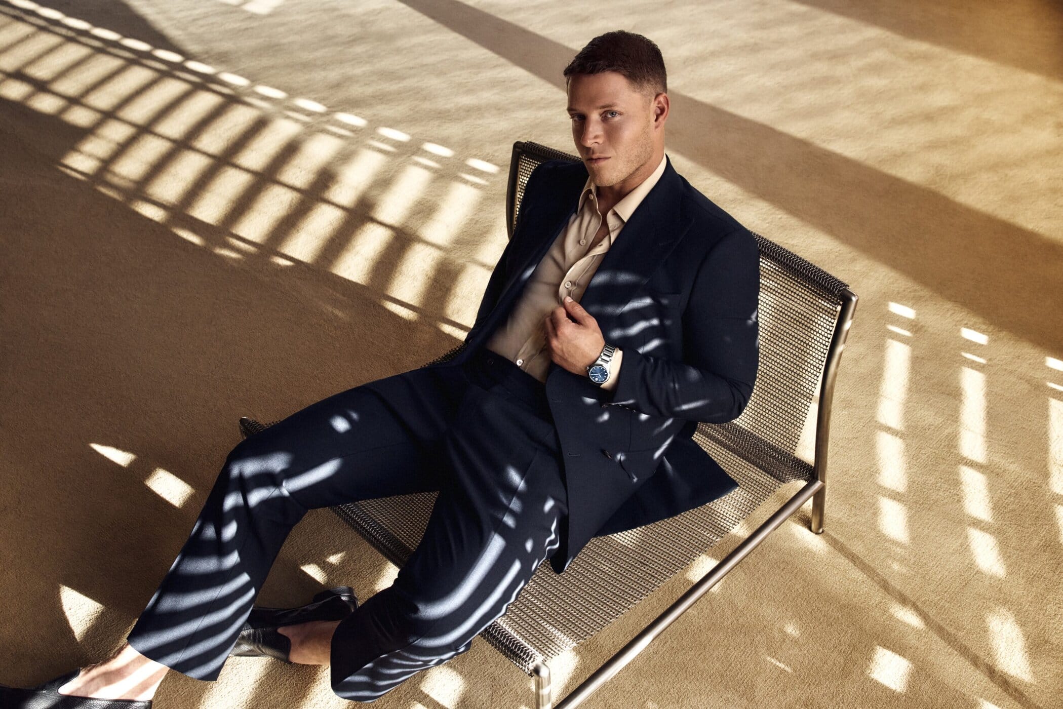 Man seated on a metal mesh bench wearing a dark suit and wristwatch in a sunlit interior space