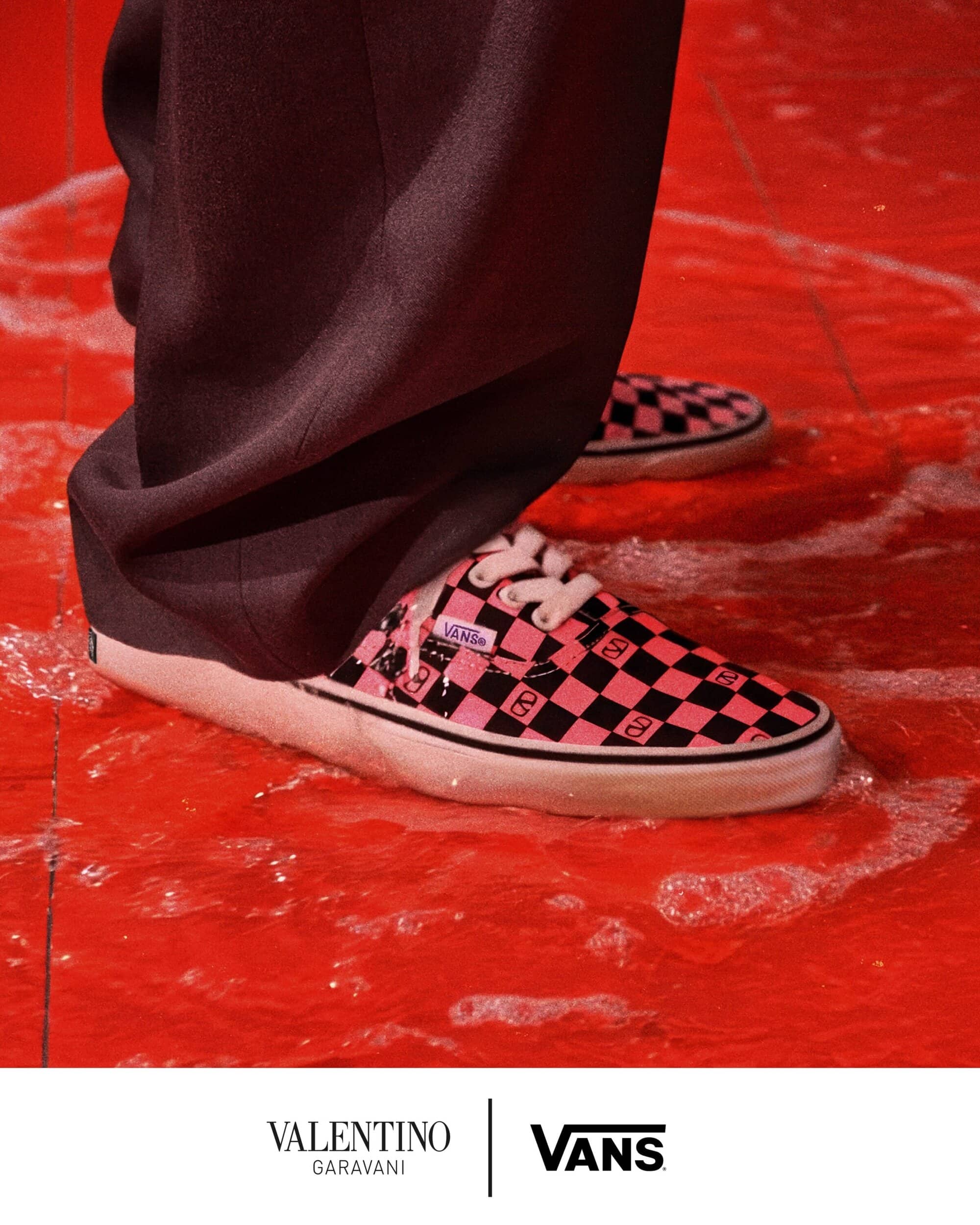 Close-up of pink and black checkered sneakers standing on a glossy red surface with white foam under dark trousers.
