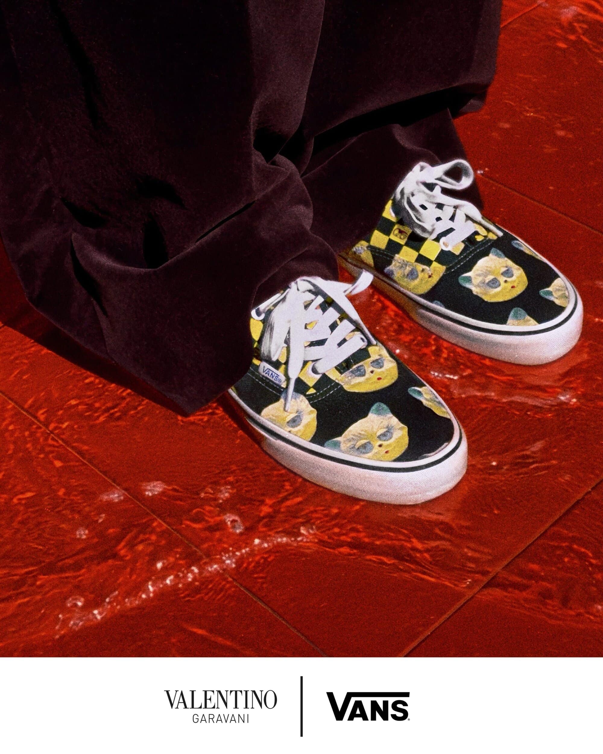 Close-up of printed sneakers and dark trousers standing on a reflective red tiled floor with brand logos at the bottom.