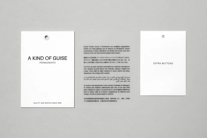 Three paper tags and labels for A Kind of Guise arrayed on a neutral grey background surface.