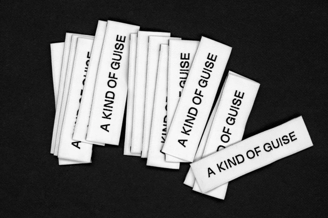 Stacked white fabric clothing labels featuring A Kind of Guise text on a dark background