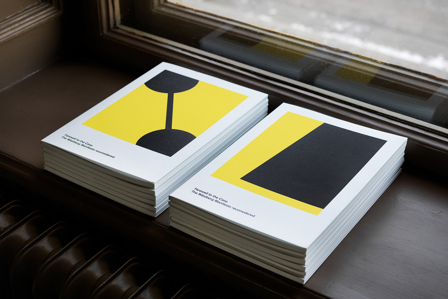Two stacks of brochures with bold yellow and black geometric cover designs resting on a wooden surface near a window