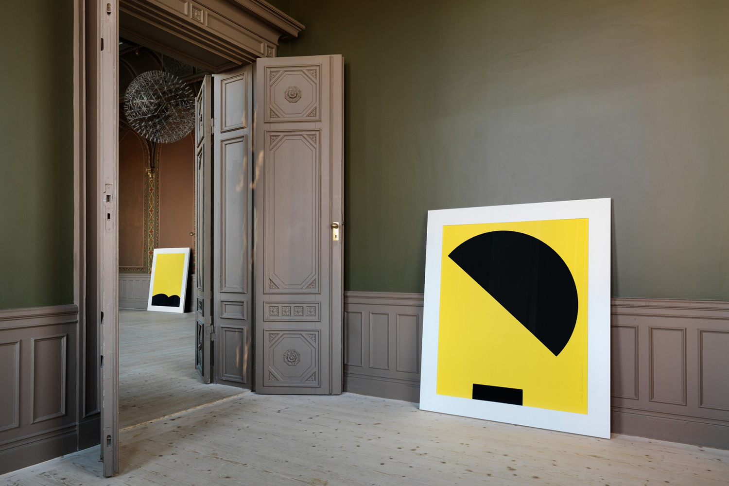 Yellow and black geometric art prints lean against panelled walls inside an elegant room with tall open double doors.