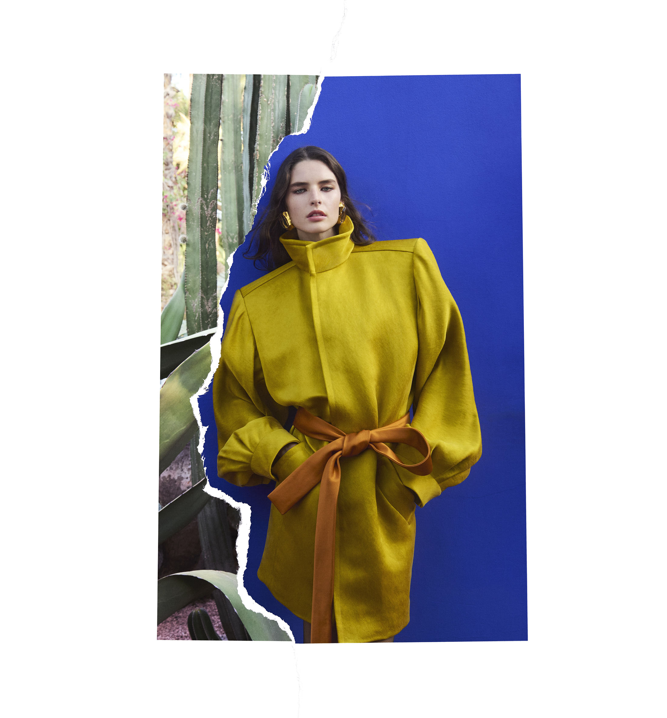 Model wearing a mustard yellow coat with a brown belt against a blue backdrop with torn paper effect