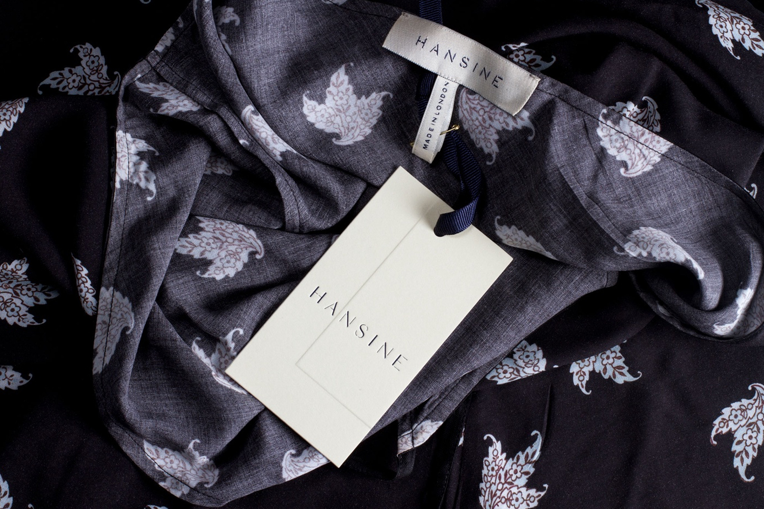 Close-up of a dark patterned garment with a white Hansine brand label and paper swing tag attached by a blue ribbon.