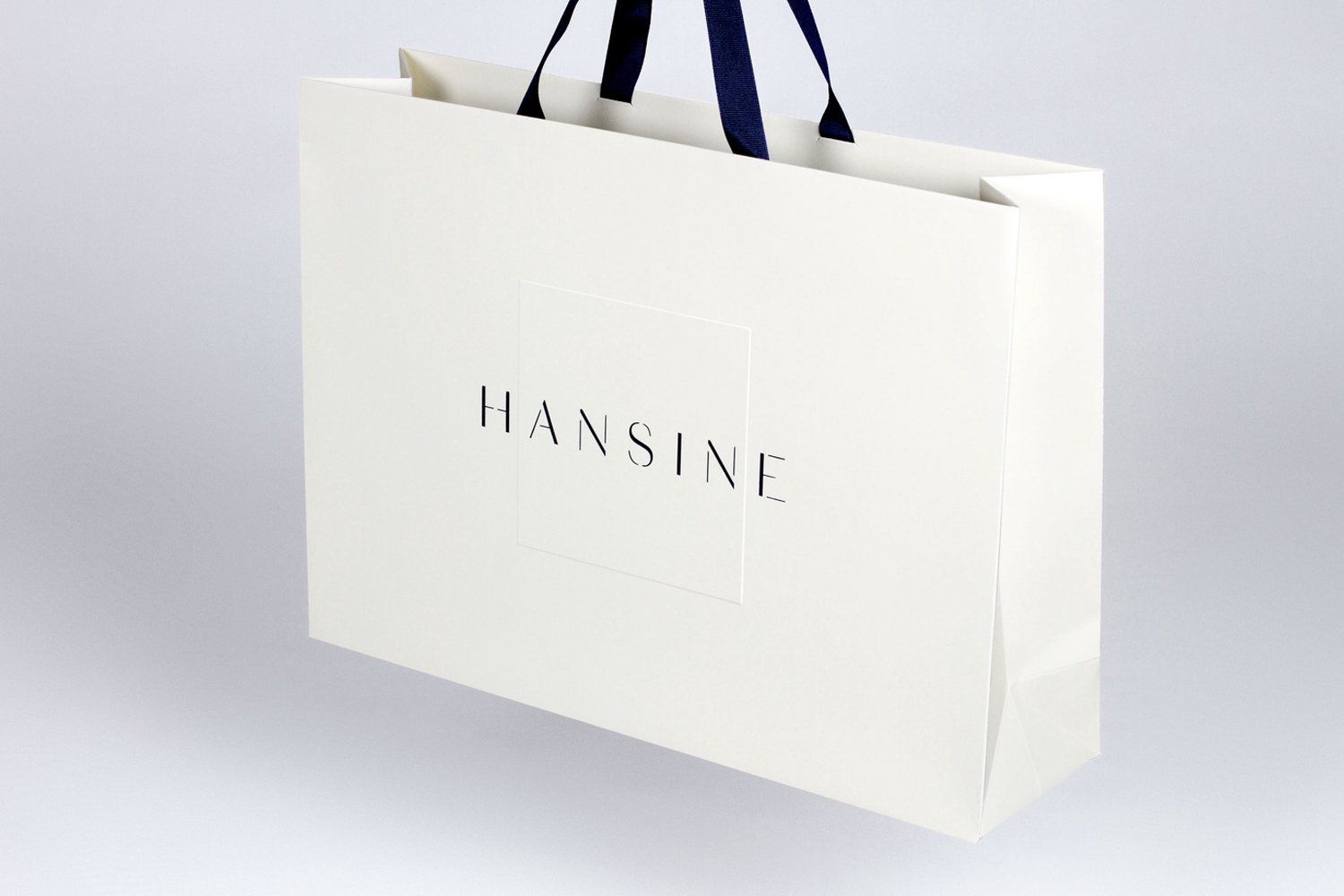 White paper shopping bag with navy ribbon handles and Hansine logo on a clean white background