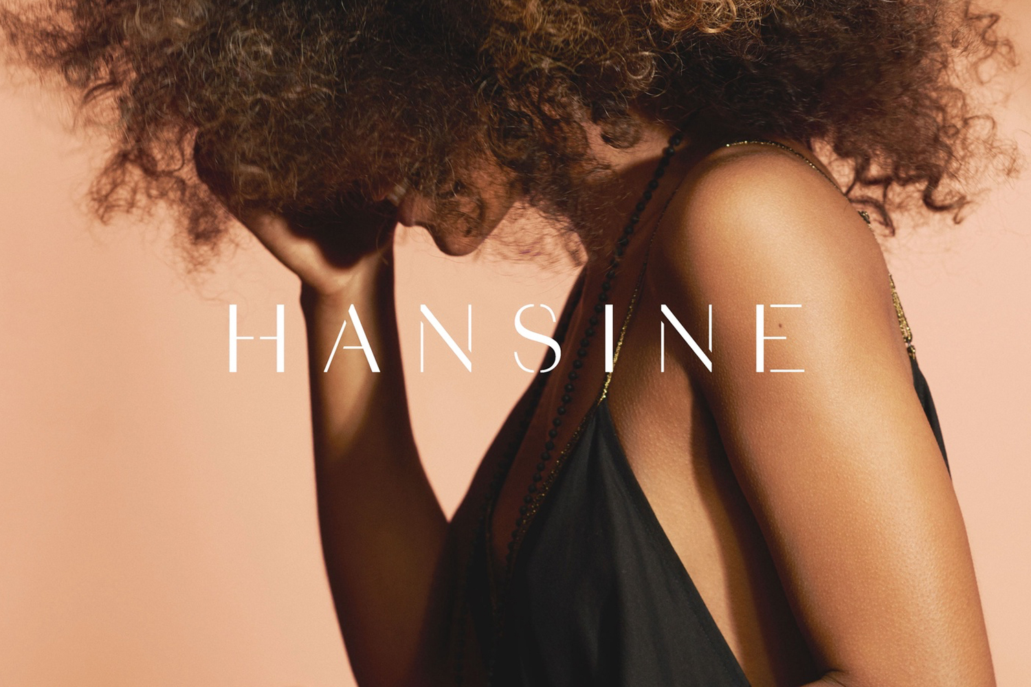 Close-up portrait of a woman with voluminous curly hair wearing a black slip dress against a warm beige background.