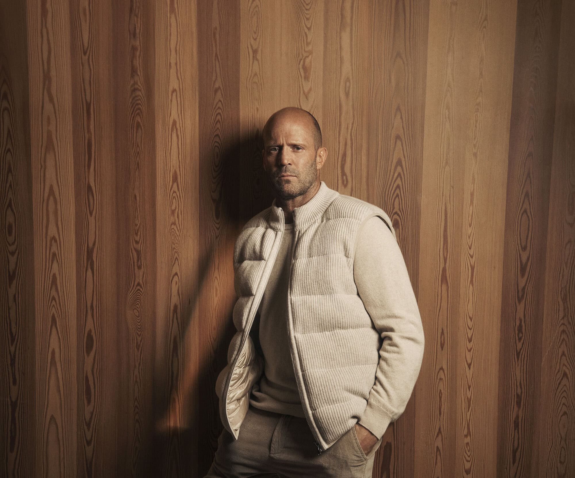 Jason Statham wearing a cream quilted vest and trousers against a vertical wood panel wall for Falconeri Fall 2025 campaign.