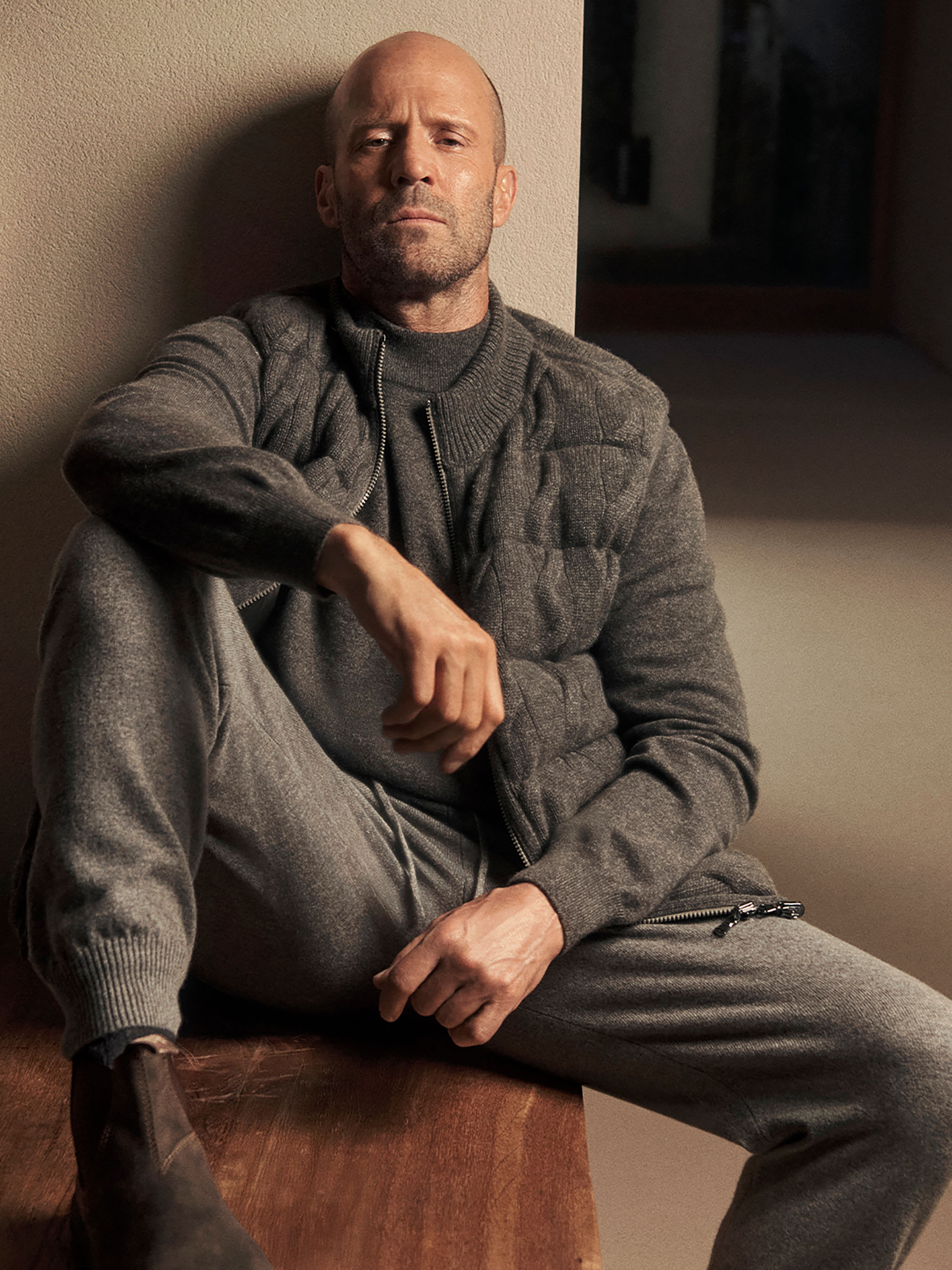 Jason Statham seated indoors wearing a grey quilted vest and matching knit trousers leaning against a wall