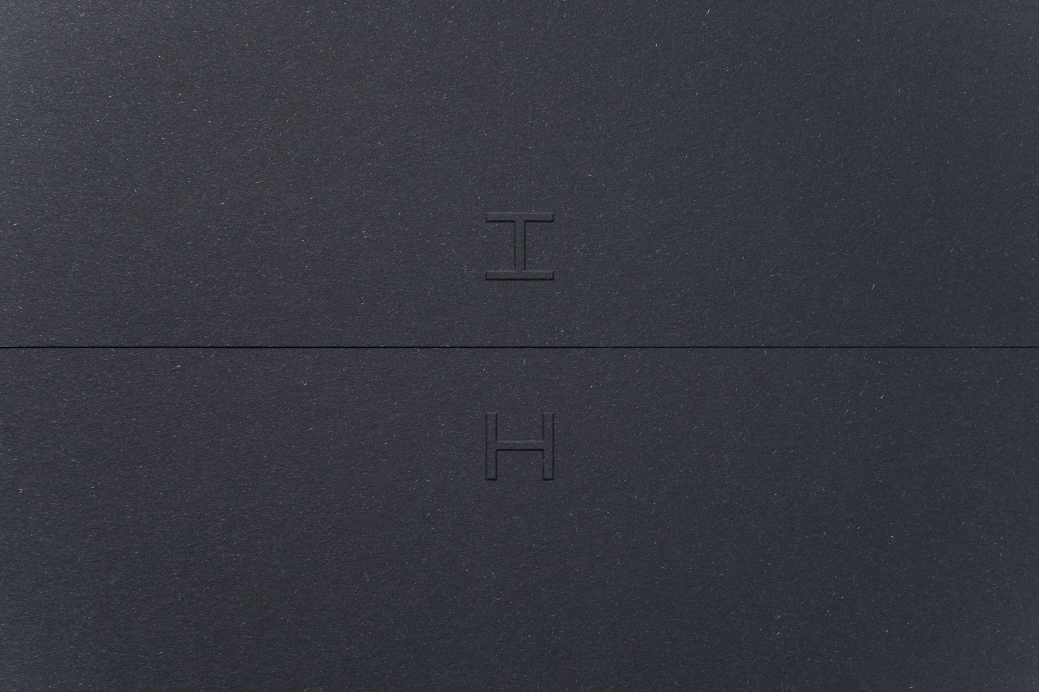 Embossed letter H monogram on dark grey textured paper surface with a horizontal seam across the center.