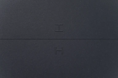 Embossed letter H monogram on dark grey textured paper surface with a horizontal seam across the center.