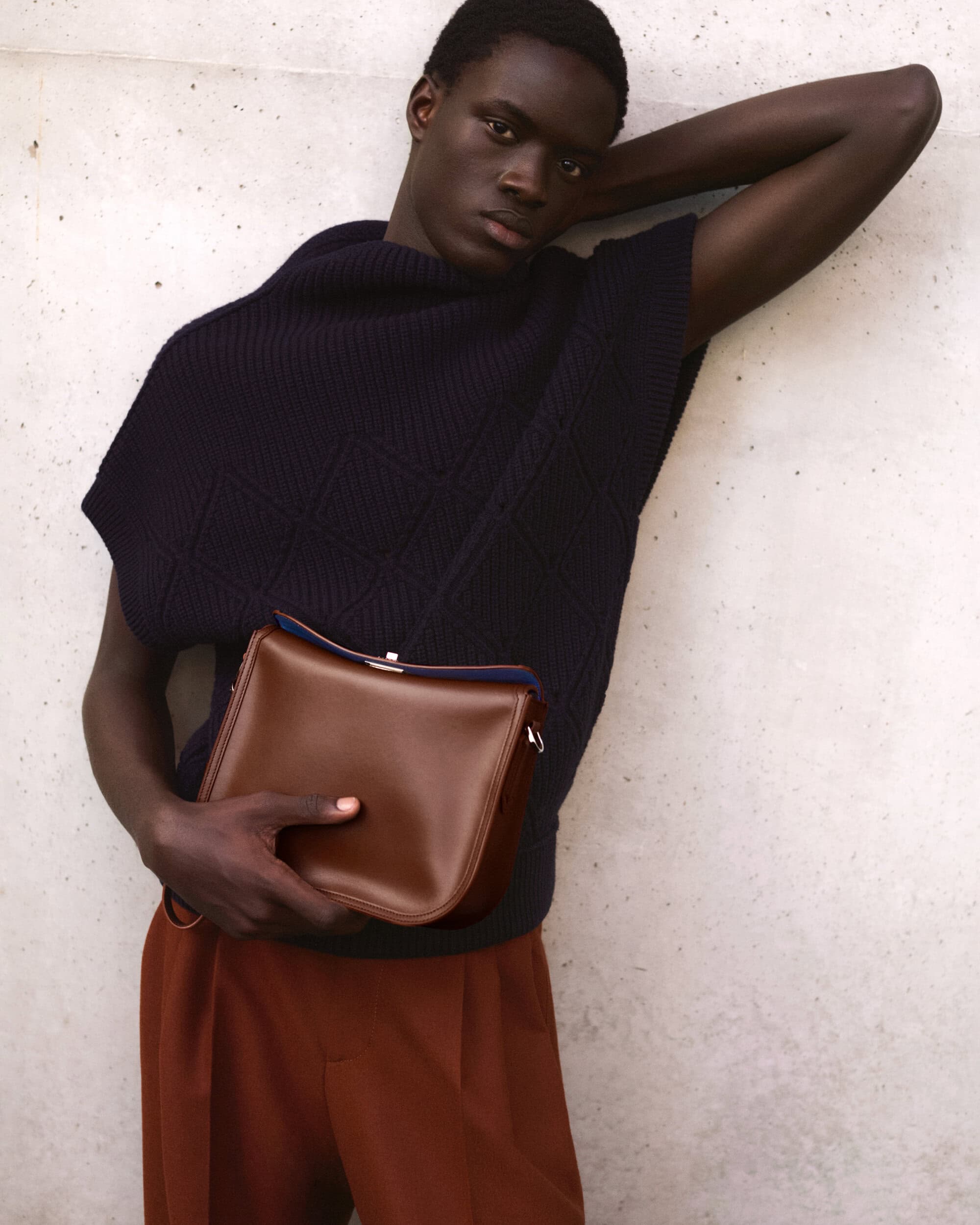 Black man wearing a dark sleeveless sweater and brown trousers holding a leather shoulder bag against a textured wall.