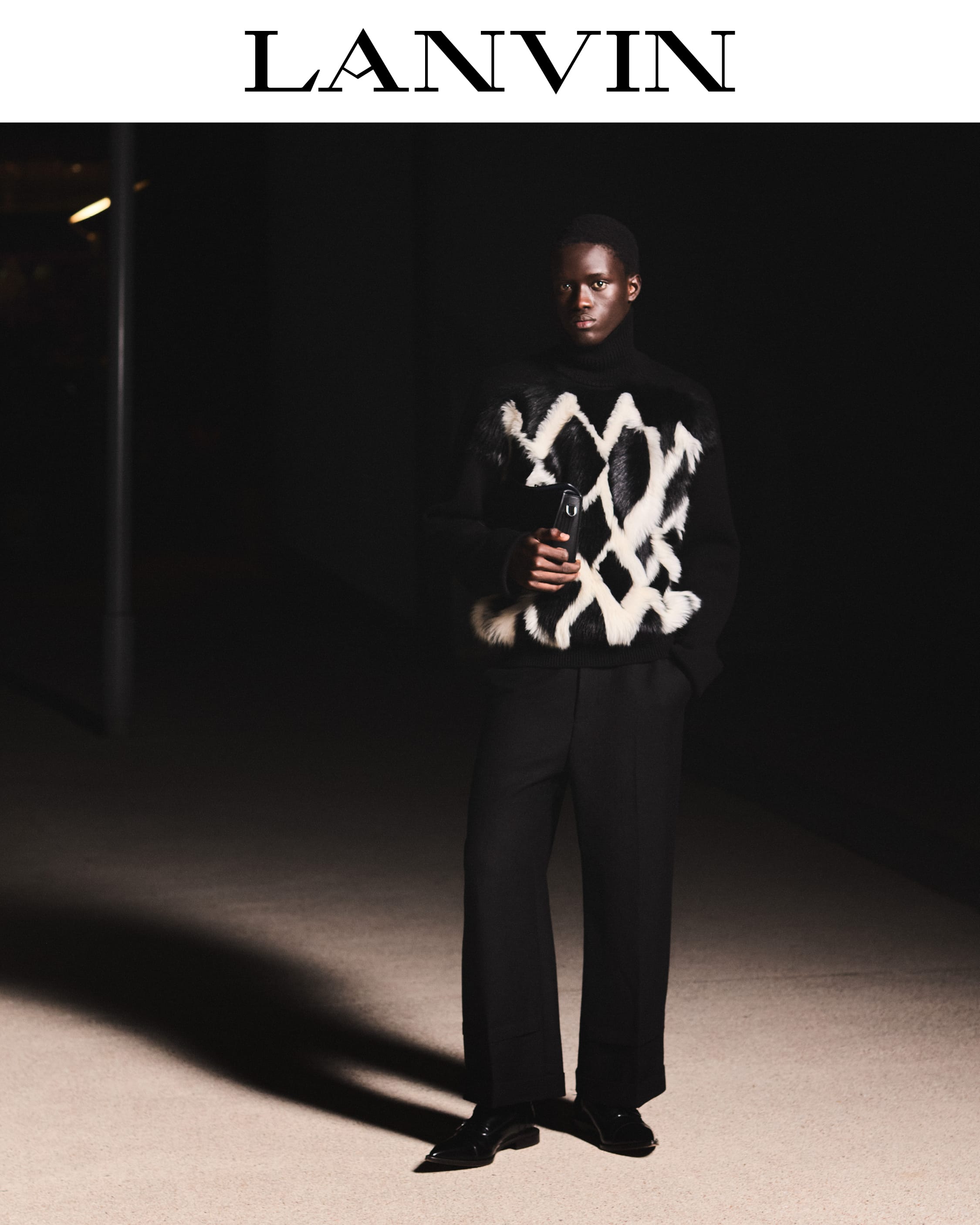 Black man wearing a patterned black sweater and dark trousers standing outdoors at night under low lighting for Lanvin