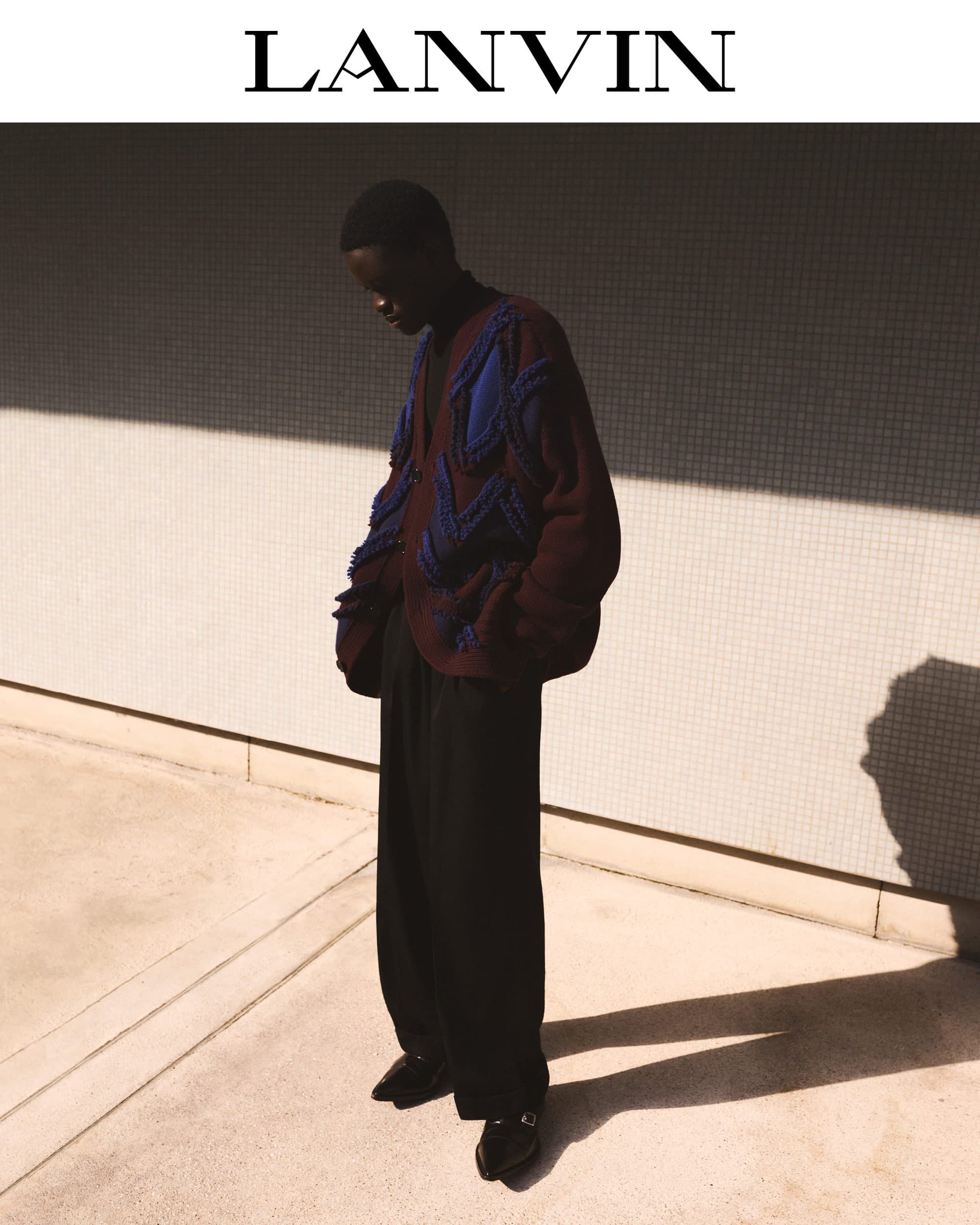 Model standing outdoors wearing a dark textured patterned cardigan and black trousers against a tiled wall