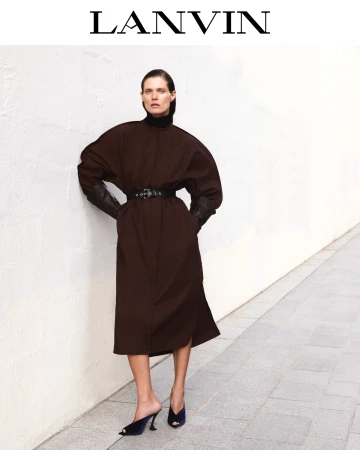 Model wearing a brown belted coat leaning against a textured white wall outdoors under natural light.