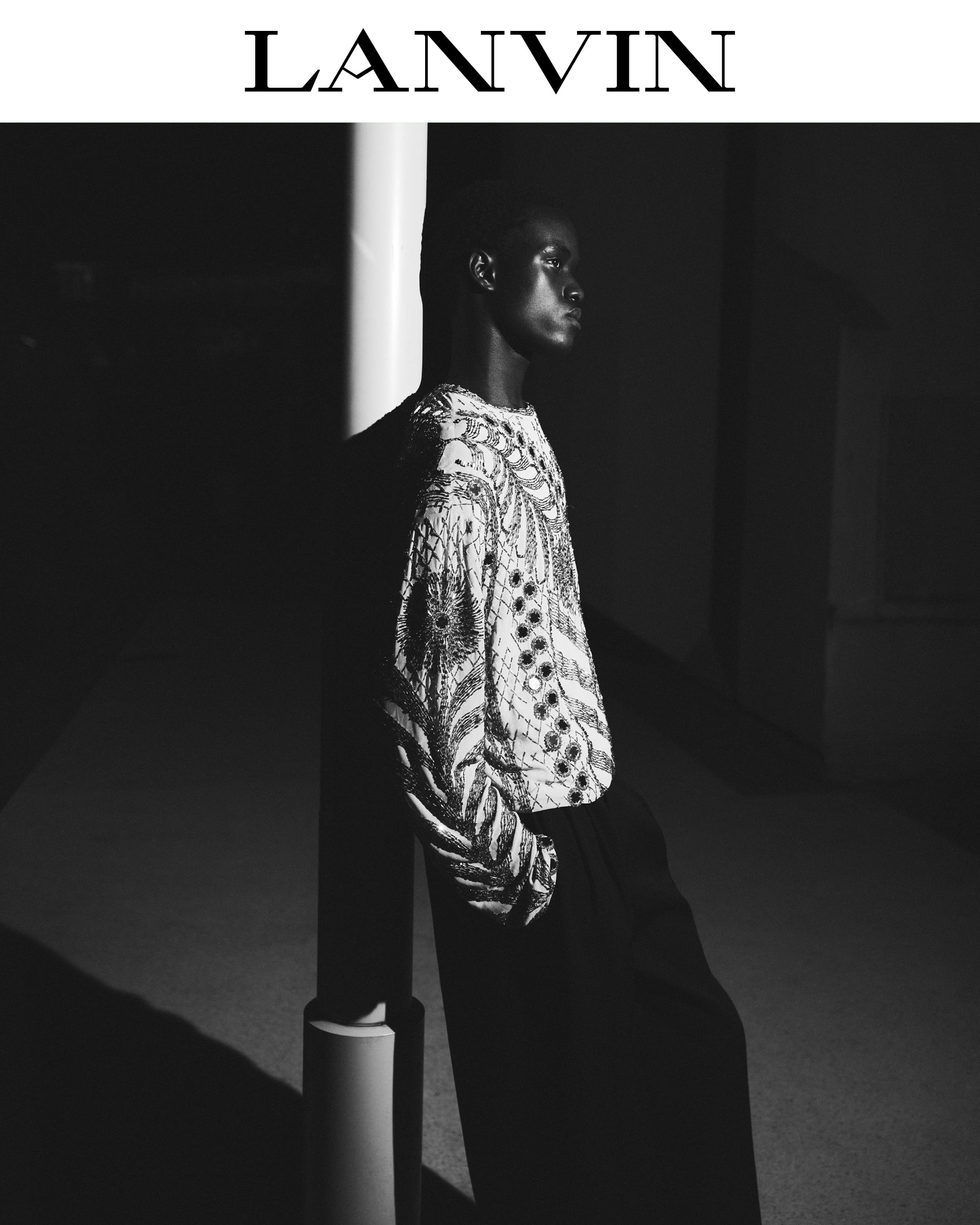 Black model wearing an embroidered patterned top leaning against a white column under low-key lighting for Lanvin Fall 2025.