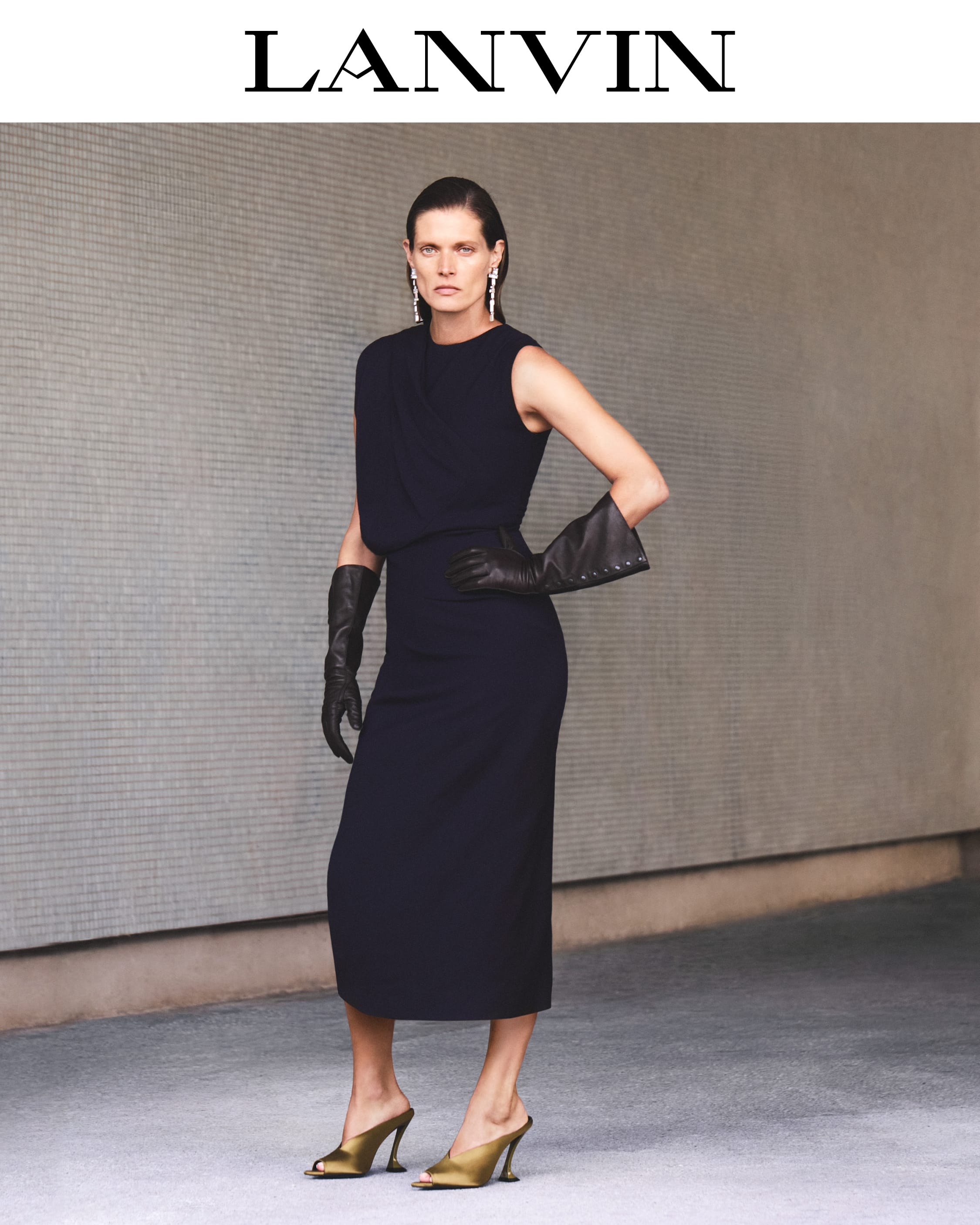 Model in dark sleeveless dress wearing long black leather gloves and gold mules against a textured wall