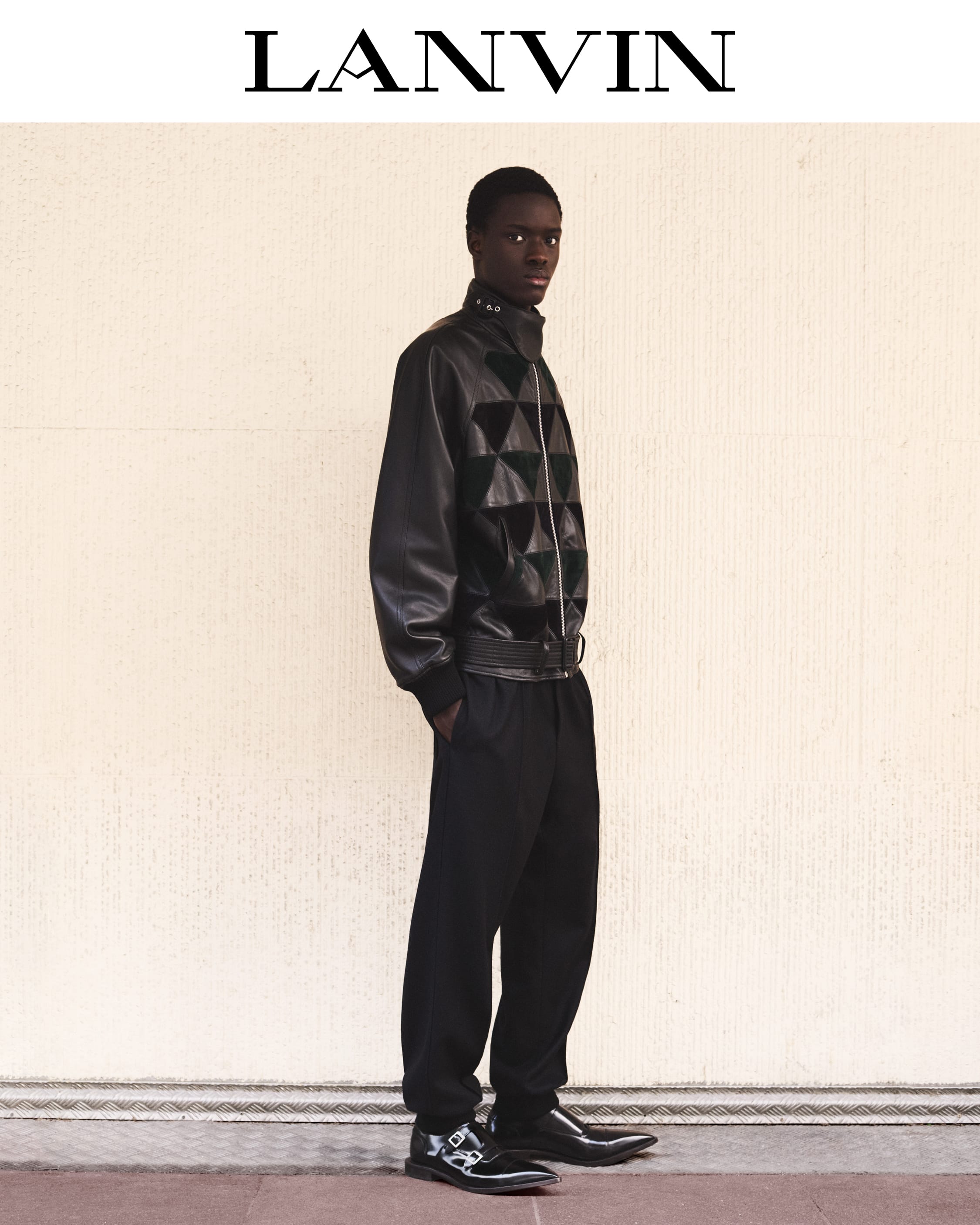 Black man wearing dark leather patterned jacket and trousers standing against light textured wall with Lanvin text above