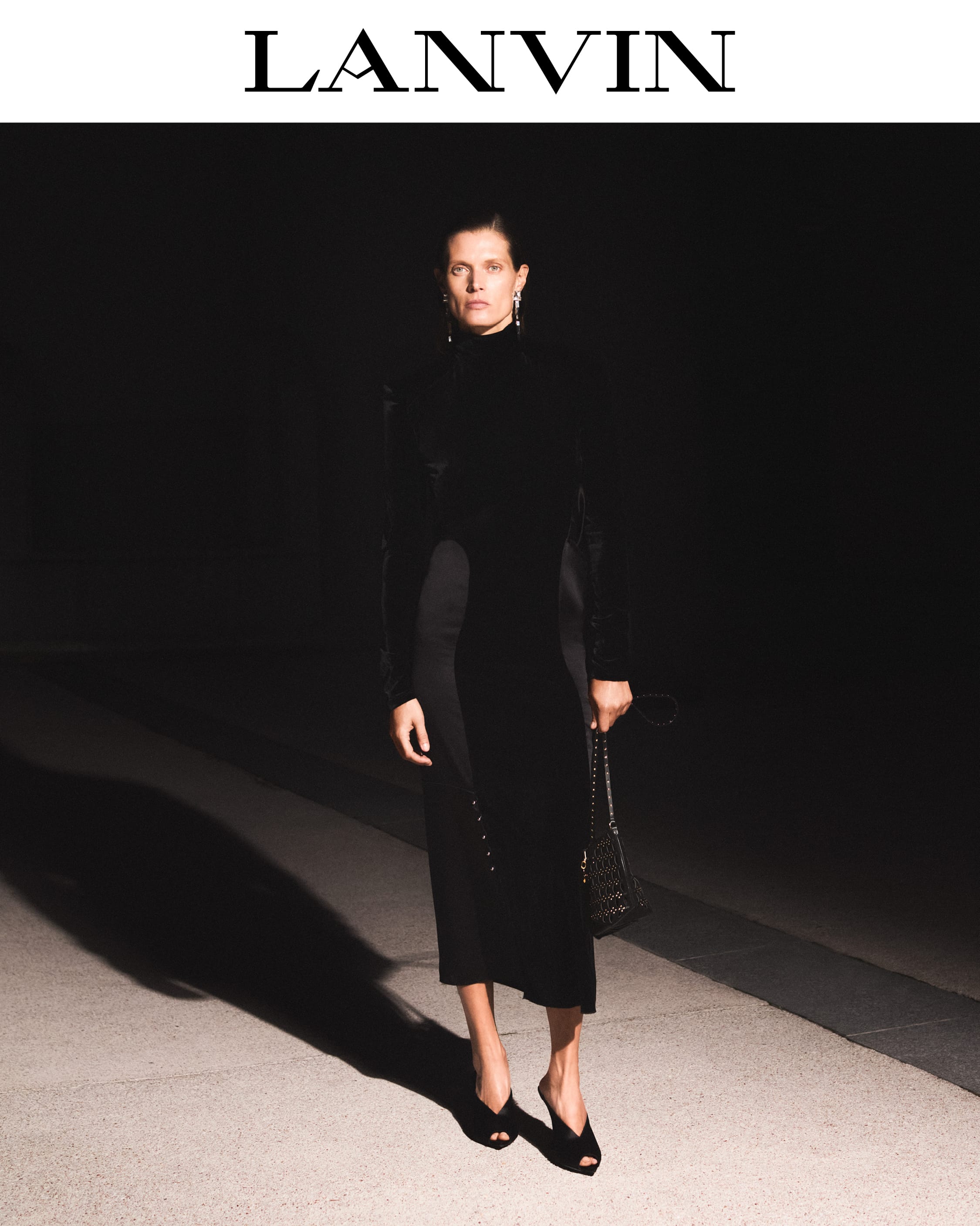 Model standing outdoors at night wearing a black dress and holding a handbag under directional street lighting