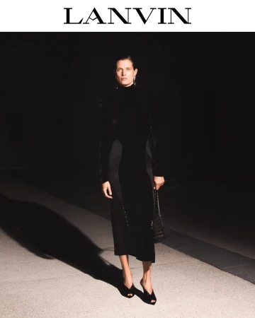 Model standing outdoors at night wearing a black dress and holding a handbag under directional street lighting