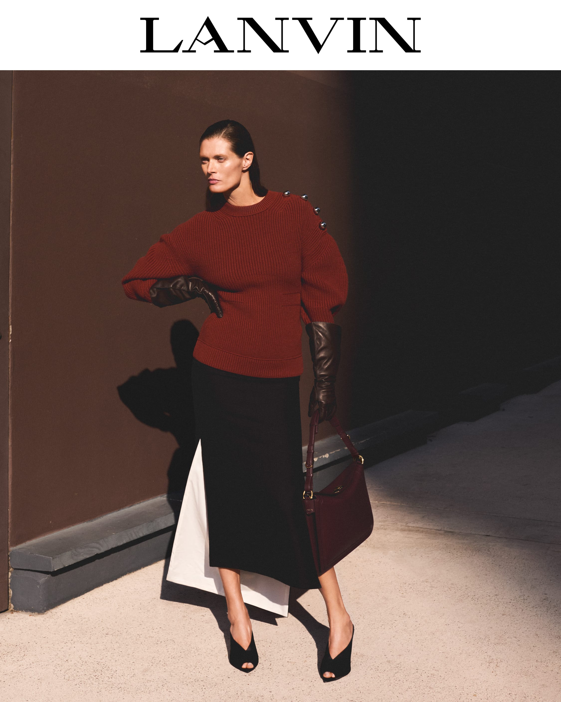 Model in a burgundy sweater and black skirt holding a handbag against a dark wall