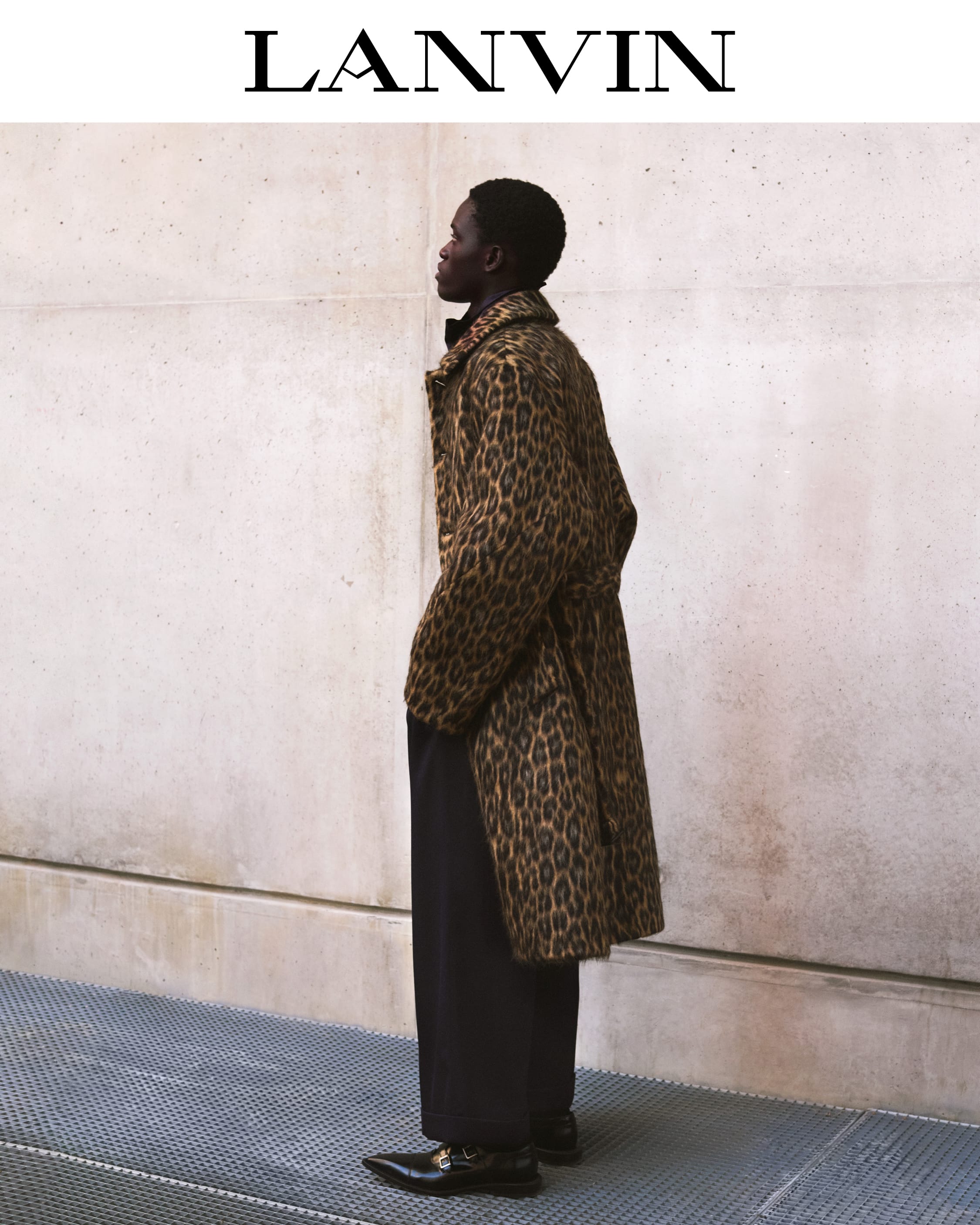 Man wearing a leopard print coat and black trousers standing against a concrete wall outdoors