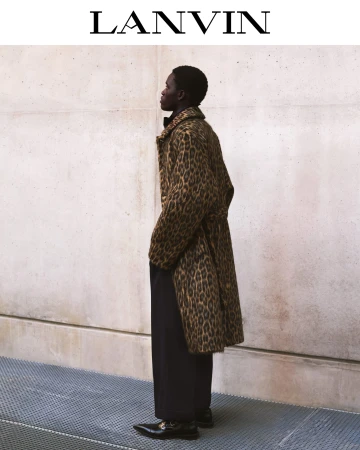 Man wearing a leopard print coat and black trousers standing against a concrete wall outdoors