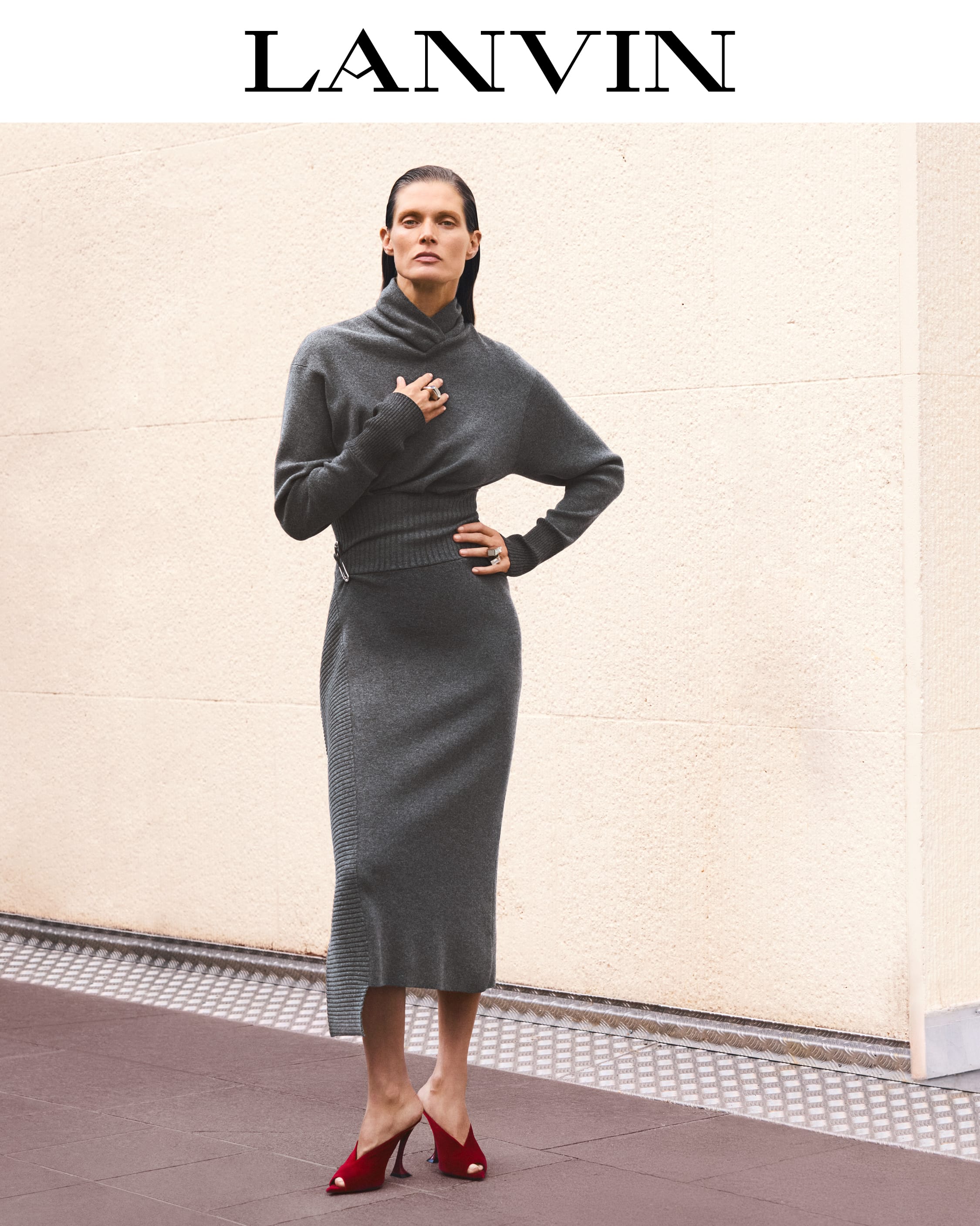 Model in a charcoal grey turtleneck knit dress and red mules posing against a textured beige wall for Lanvin Fall 2025.
