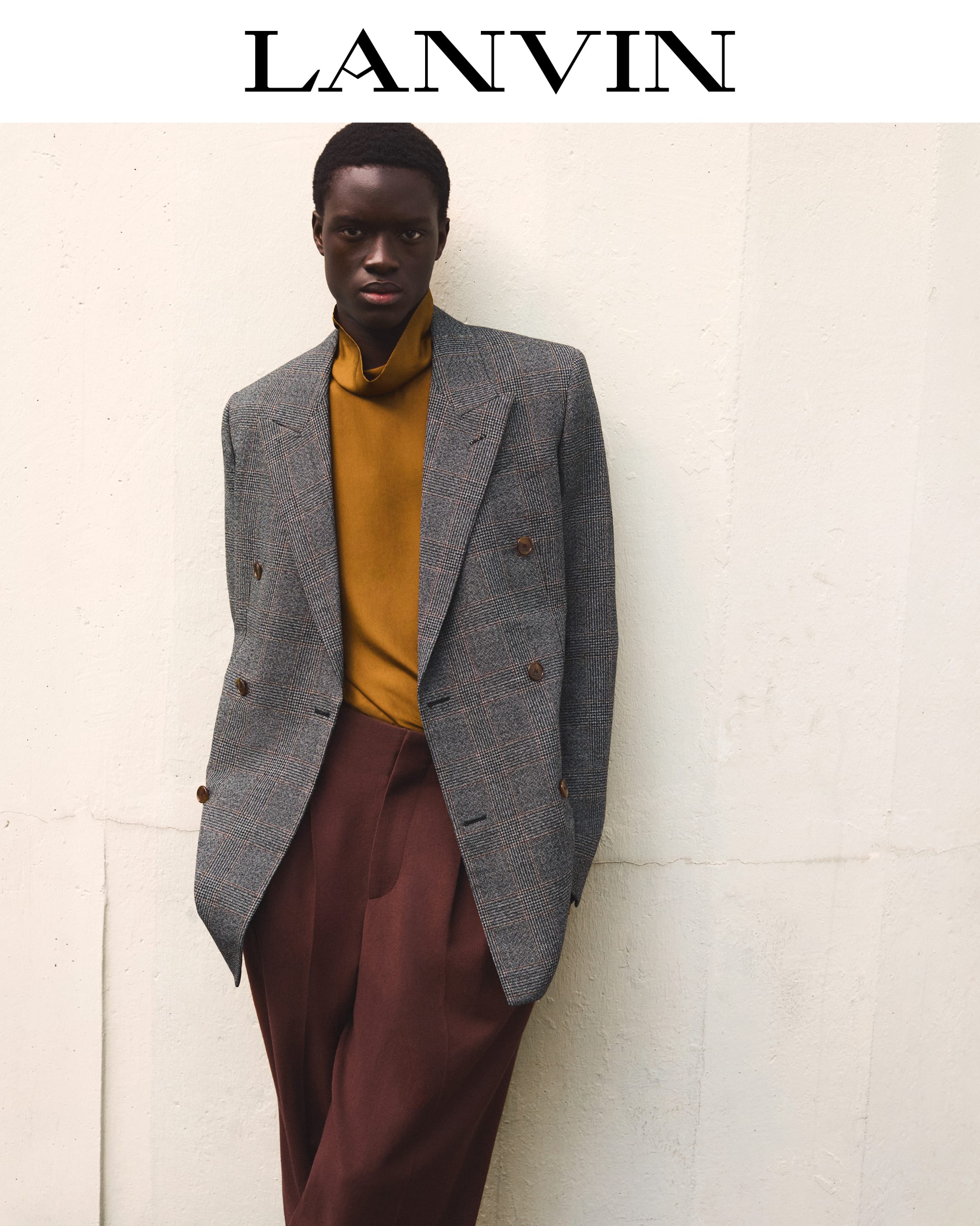 Black man wearing a grey tweed double-breasted blazer, mustard turtleneck, and brown trousers against a plain white wall.