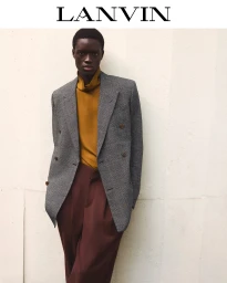 Black man wearing a grey tweed double-breasted blazer, mustard turtleneck, and brown trousers against a plain white wall.