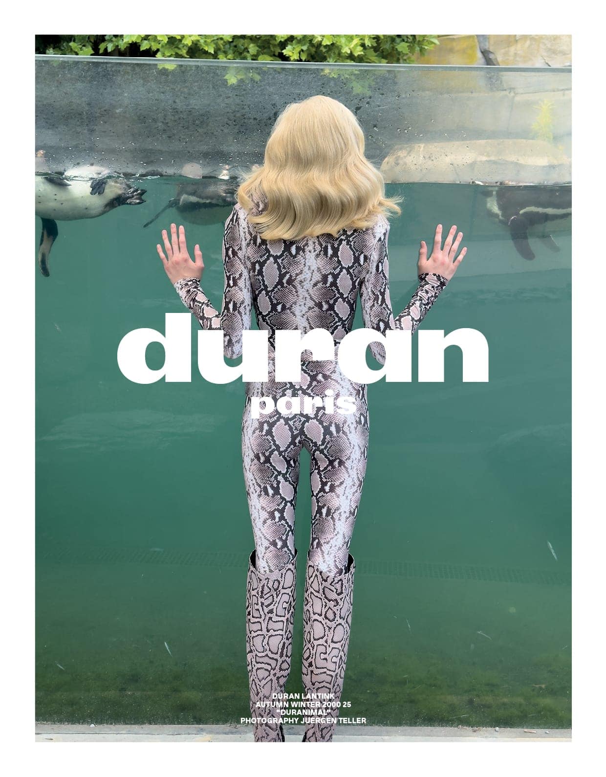 Model in a snakeskin print bodysuit with matching boots standing before a large glass aquarium tank with penguins swimming