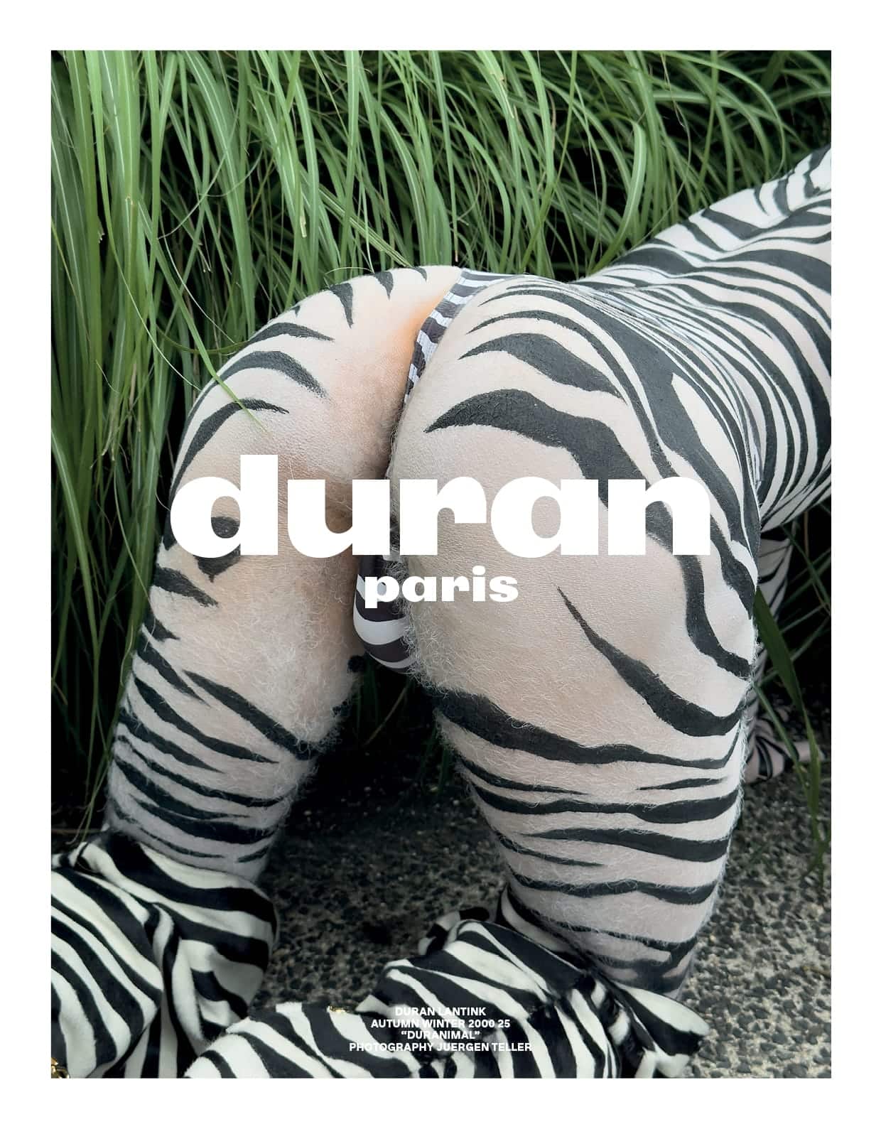 Model wearing zebra-patterned body paint and apparel posing on all fours amidst tall green grass for Duran Lantink DURANIMAL campaign