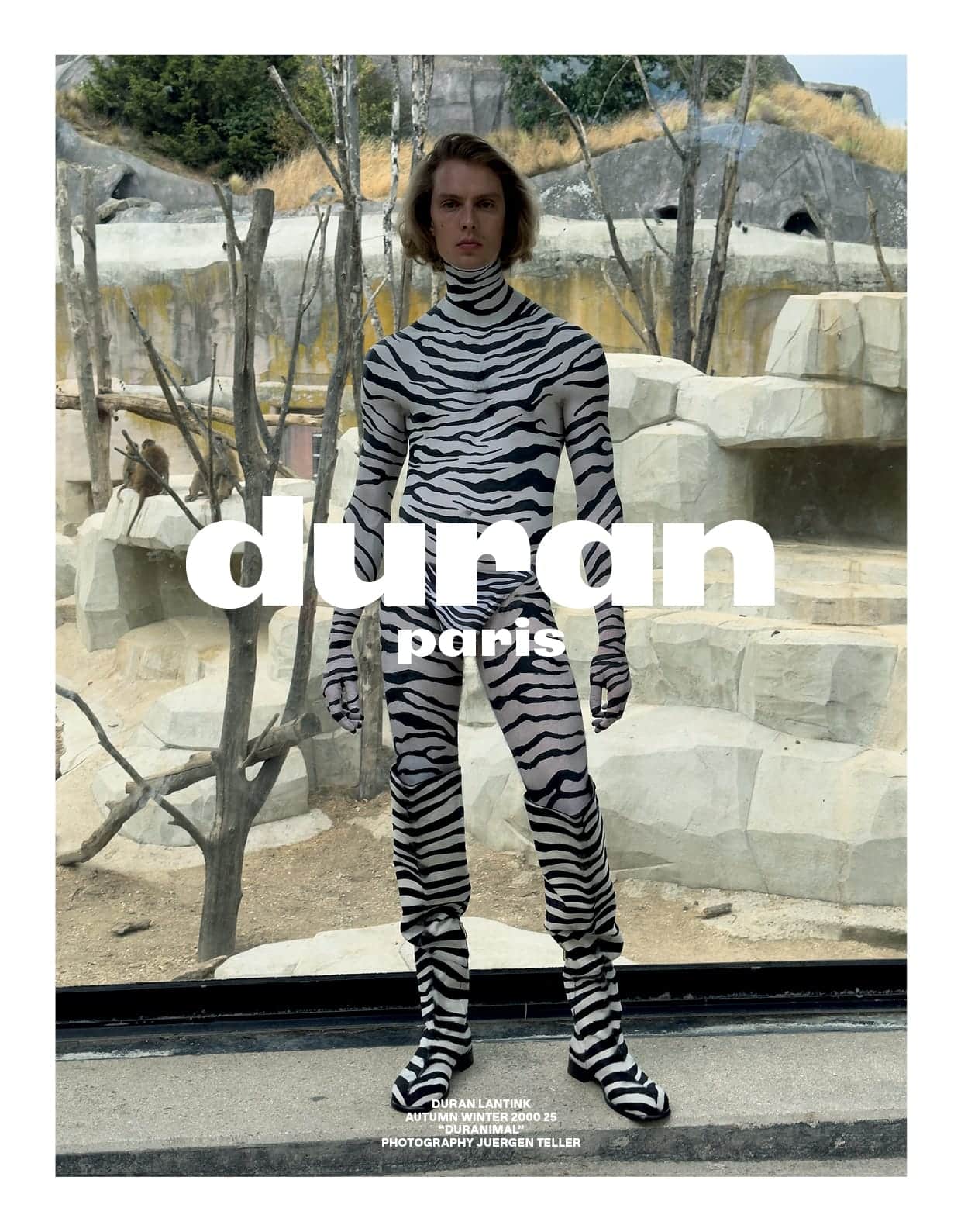 Model in zebra-print bodysuit standing in front of an enclosure with rock formations and bare trees