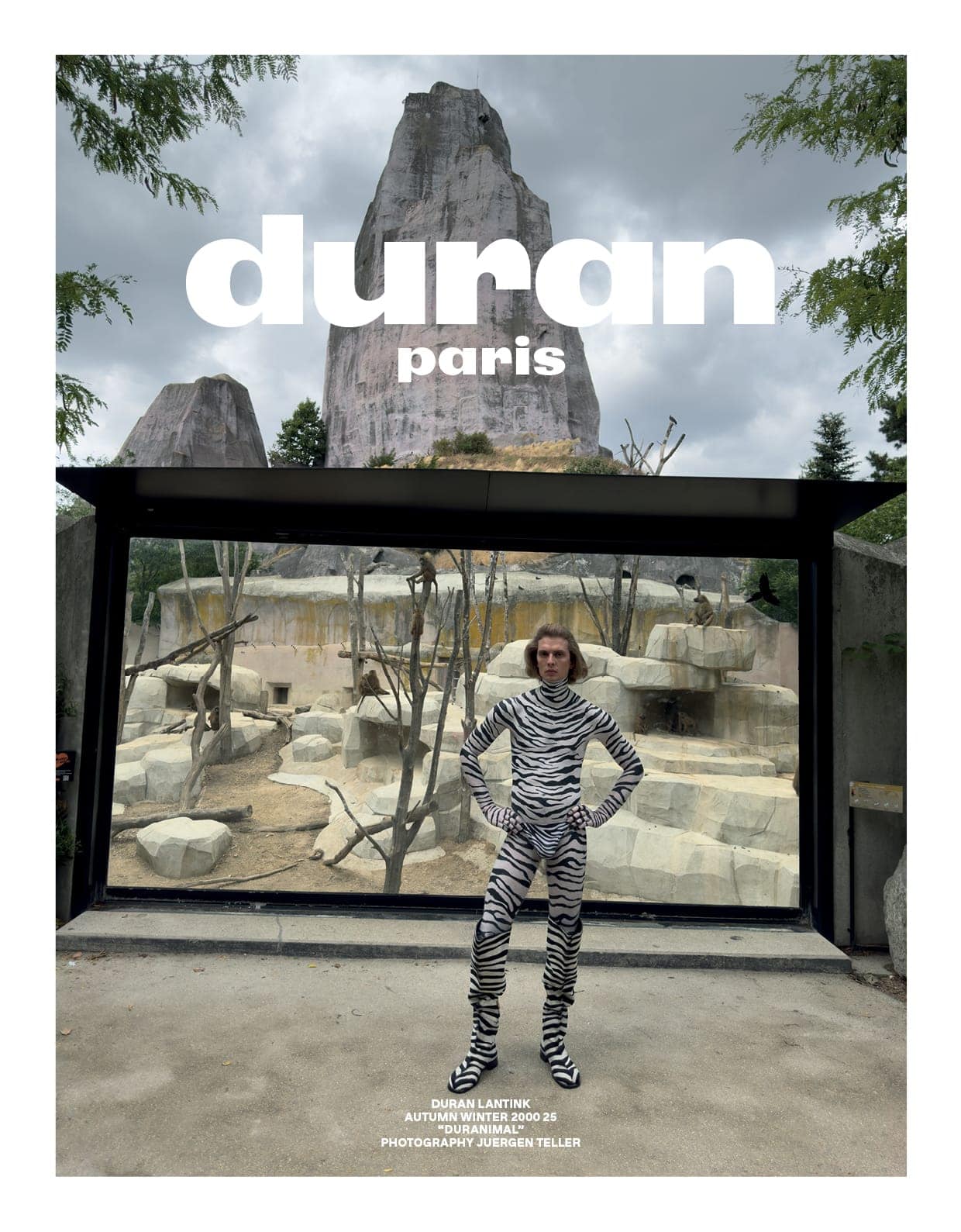 Model in zebra-print patterned bodysuit standing in front of an animal enclosure enclosure at a zoo under an overcast sky.
