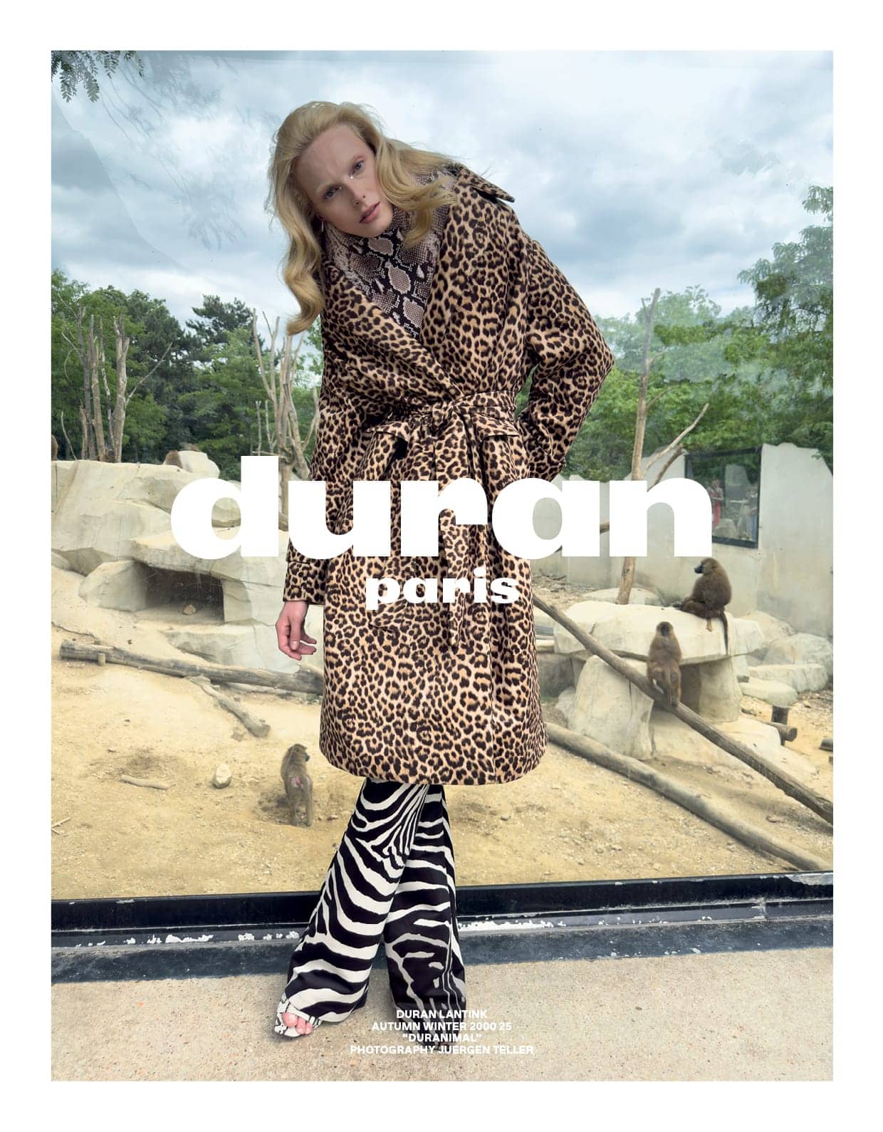 Model wearing a leopard print coat and zebra print trousers posing in front of an outdoor animal enclosure.