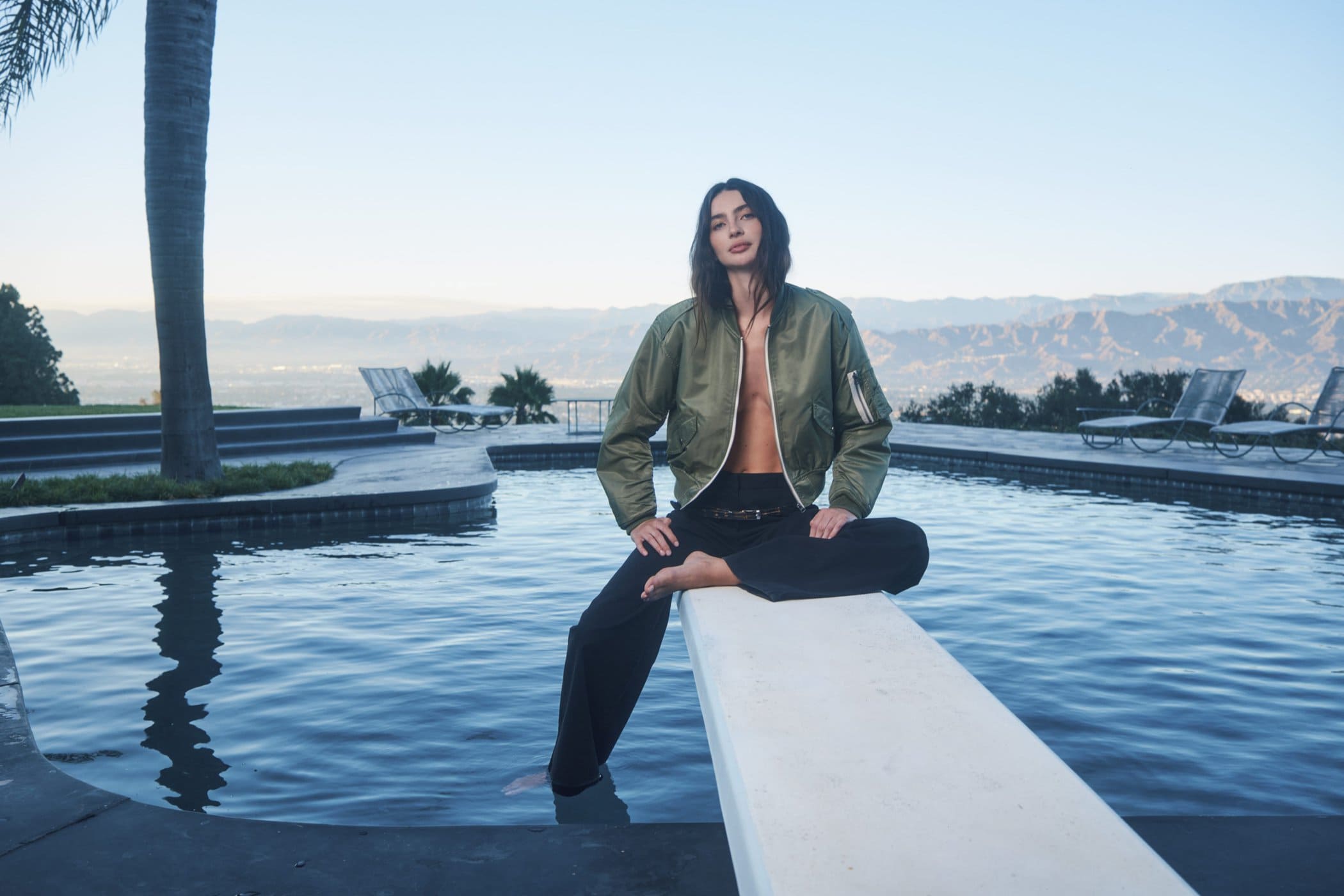 Model seated on a diving board wearing an olive green bomber jacket and dark trousers by a swimming pool
