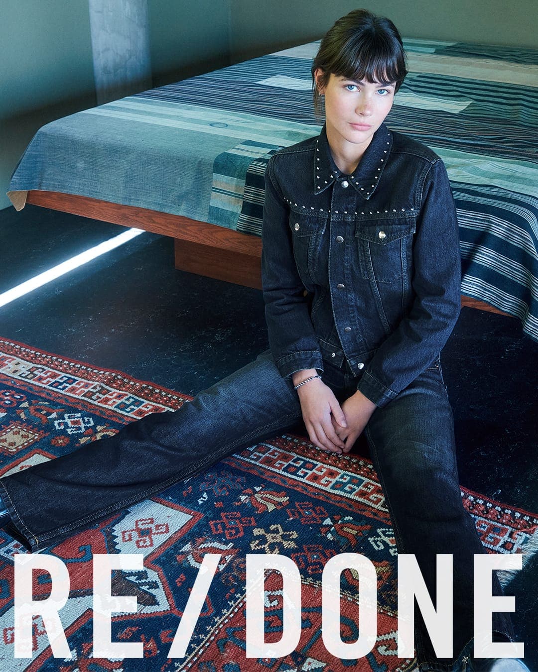 A model seated on an patterned rug in front of a made bed wearing a dark denim jacket with silver studs and matching jeans.