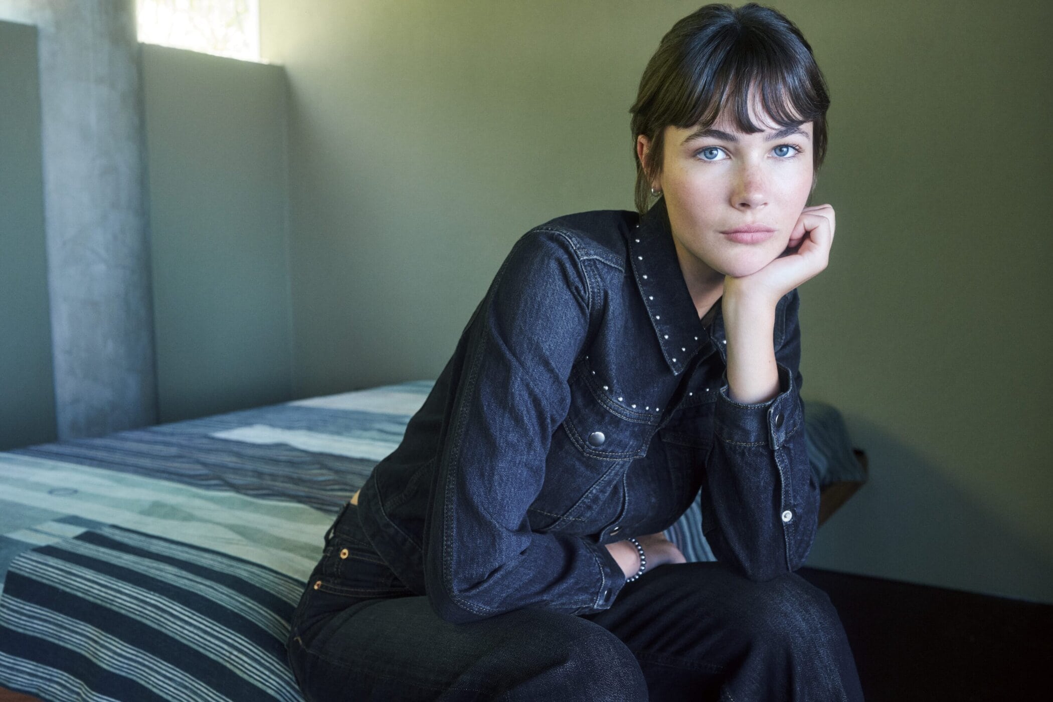 Woman in dark denim jacket and pants resting chin on hand inside a bedroom