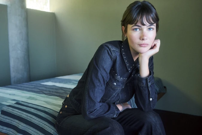 Woman in dark denim jacket and pants resting chin on hand inside a bedroom