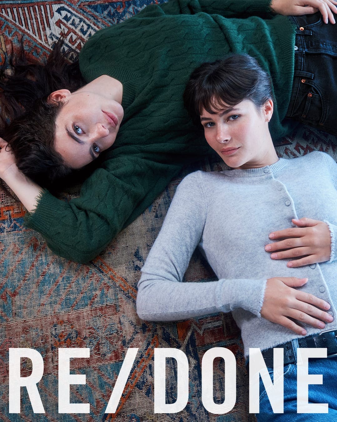Two women lying on an ornate patterned rug wearing knit sweaters and denim jeans for RE/DONE