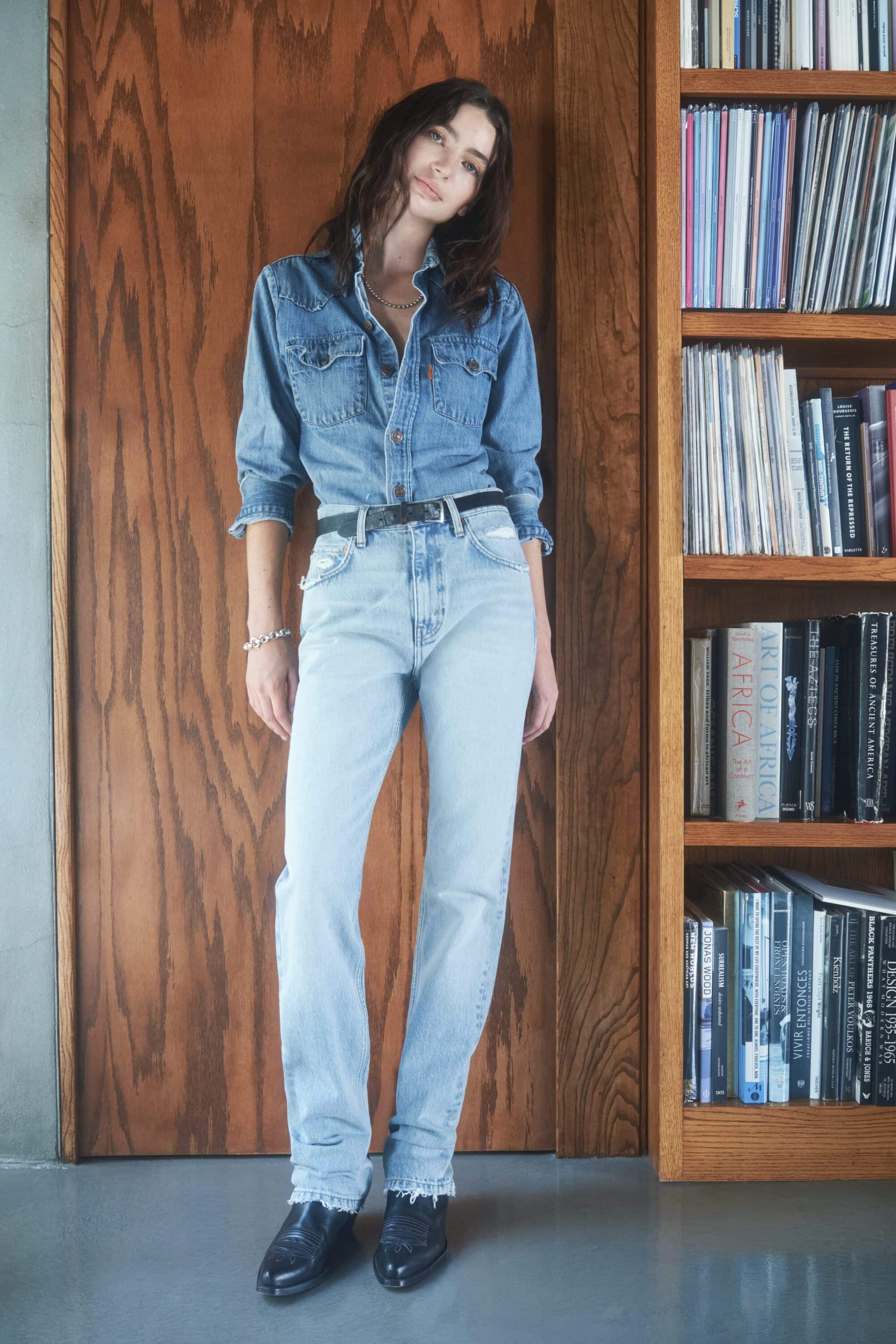 Model wearing a blue denim shirt and light wash jeans standing indoors next to a tall wooden bookshelf.