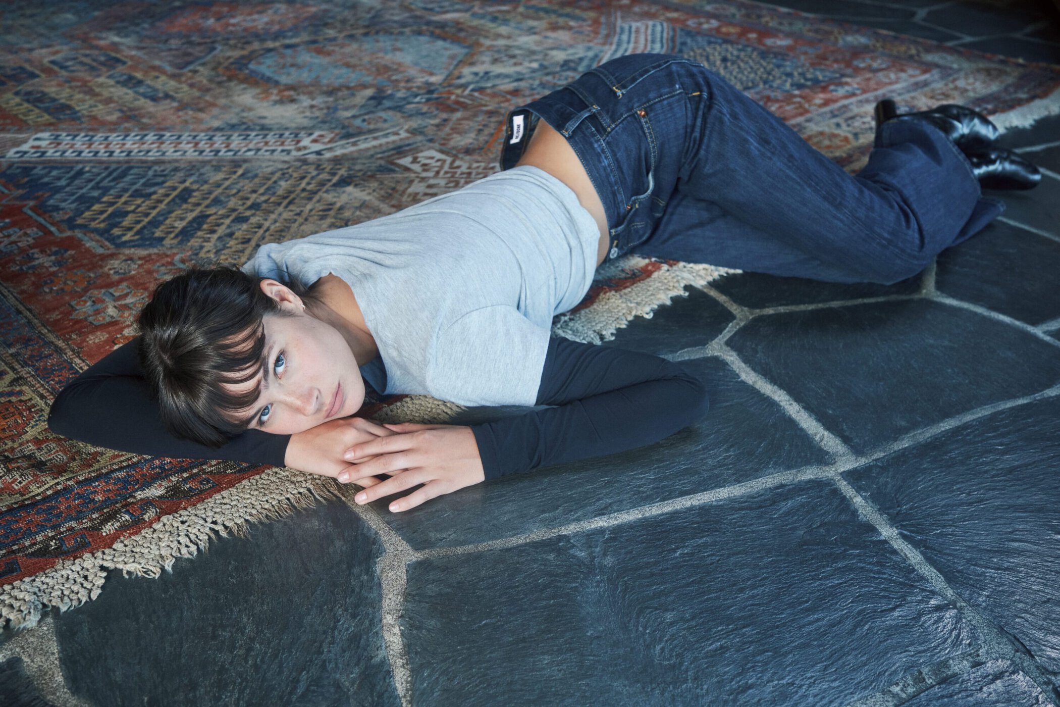 Model lying on a patterned rug and stone floor wearing dark denim jeans and a layered grey top