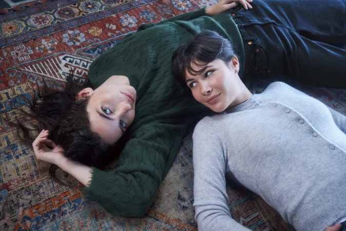 Two women lying on an ornate patterned rug wearing a green cable knit sweater and a grey cardigan for RE/DONE Fall 2025 ad campaign
