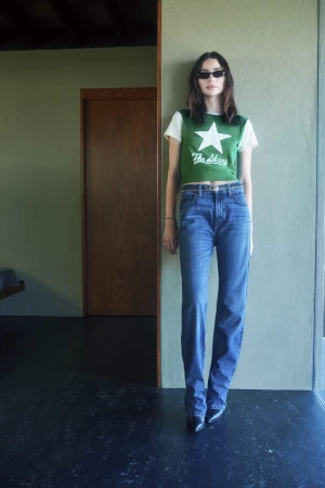 Model in dark sunglasses and green star t-shirt leaning against a wall indoors for RE/DONE