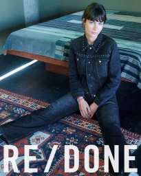 A model seated on an patterned rug in front of a made bed wearing a dark denim jacket with silver studs and matching jeans.