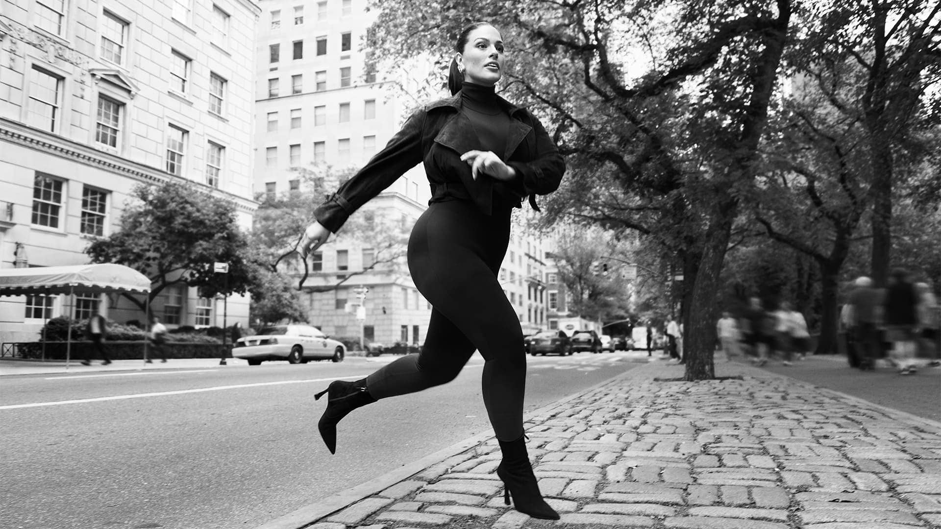 Ashley Graham running along a New York street wearing a dark jacket and heels in a black and white campaign photograph.